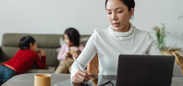 Woman in white turtleneck sitting at her kitchen table writing something down with laptop in front of her. Her two children play int the background.